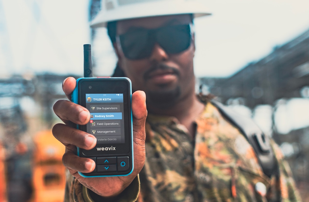Cover image for a blog on how to build a better frontline employee experience with smart radios. A frontline worker wearing sunglasses and a hard helmet holds up a Walt Smart Radio, with a close-up on all the different channels in their organization.