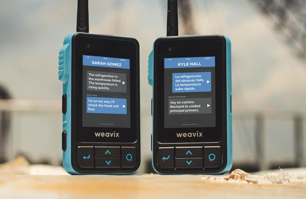 Two Walt Smart Radios, which are real-time translation devices, on a manufacturing site. These two Walts display AI translations of incoming messages as English and Spanish transcriptions.